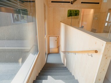 Modernes Treppenhaus mit Holzverkleidung an Wänden und Decke, Holzhandlauf und großem Fenster mit Außenblick. | © LibertydotHome GmbH