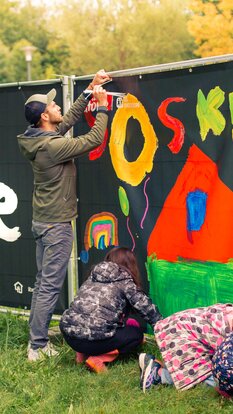 Kinder und ein Erwachsener bemalen gemeinsam einen Zaun mit buntem Wandbild | SOS Kinderdorf  | © LibertydotHome GmbH