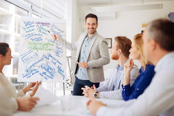 Mann erklärt Whiteboard Mann deutet vor Publikum mit einem Stift auf ein beschriebenes Whiteboard