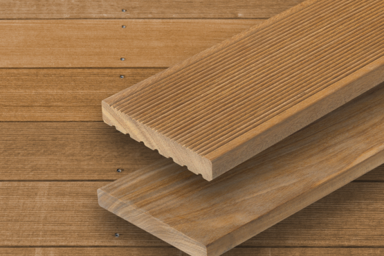 Terrassendielen aus Holz Zwei braune parallele Terrassendielen mit sichtbarer Maserung und Riffelung liegen auf einer Fläche aus ähnlichem Holz mit horizontalen Brettern im Hintergrund.