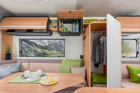 Blick in einen Camper-Van mit Holz-Tisch, gedecktem Essbereich, offenen Staufächern, Kleiderschrank und Aussicht auf Berge und Natur durch die Fenster. | © Knaus Tabbert