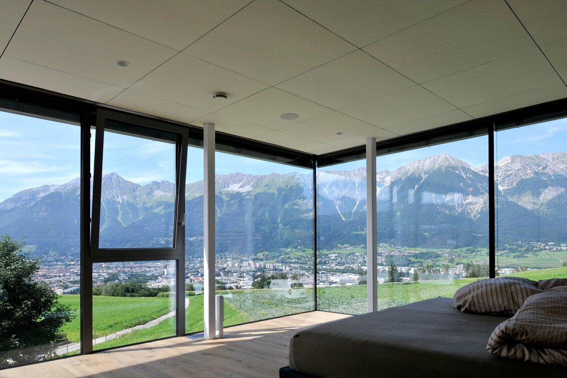 Modernes Schlafzimmer mit Panoramablick auf die Berge | © Whisperwool Schlafzimmer mit hellem Holzfußboden, großflächigen Fenstern und Blick auf grüne Berglandschaft – minimalistisch eingerichtet mit Fokus auf die Aussicht. | © Whisperwool