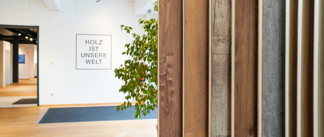 Moderner Innenraum mit Holzfußboden und mehreren Bodenmustern, die senkrecht zur Ansicht präsentiert werden; im Hintergrund is auf der Wand  „HOLZ IST UNSERE WELT“ geschrieben und eine dekorative Pflanze.