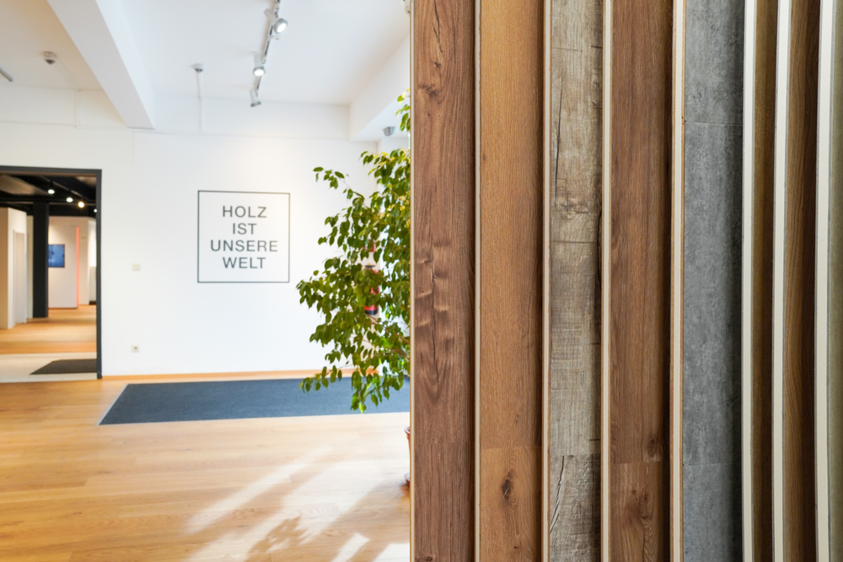 Ausstellung von Bodenmustern im Schauraum Moderner Innenraum mit Holzfußboden und mehreren Bodenmustern, die senkrecht zur Ansicht präsentiert werden; im Hintergrund is auf der Wand „HOLZ IST UNSERE WELT“ geschrieben und eine dekorative Pflanze.