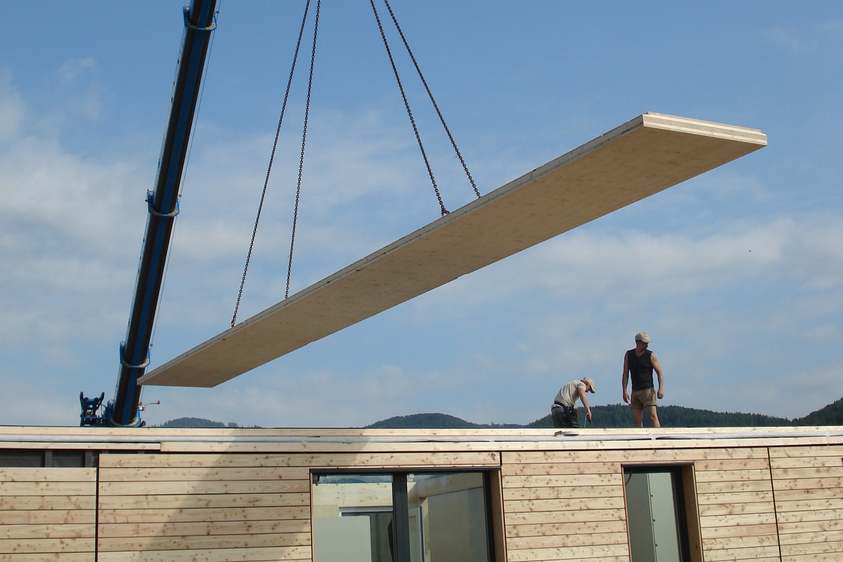 Holzbau mit Kranmontage Auf einer Baustelle wird ein großes Holzpaneel per Kran auf das Dach eines Gebäudes gehoben; zwei Arbeiter positionieren es, im Hintergrund Hügel und bewölkter Himmel.