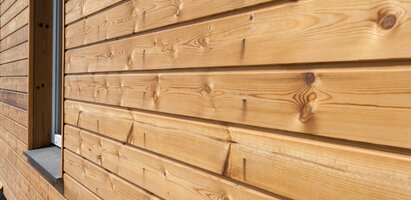 Die Aufnahme zeigt eine Fassade aus horizontal verlegten Holzplanken mit sichtbarer Maserung und Astlöchern. Links ist ein Teil eines Fensterrahmens zu erkennen.