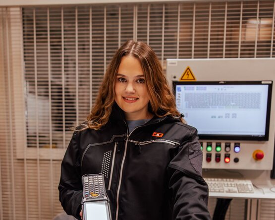 Person in schwarzer Jacke hält einen Scanner mit Tasten und Display, im Hintergrund ein beleuchtetes Bedienfeld mit Knöpfen und Bildschirm